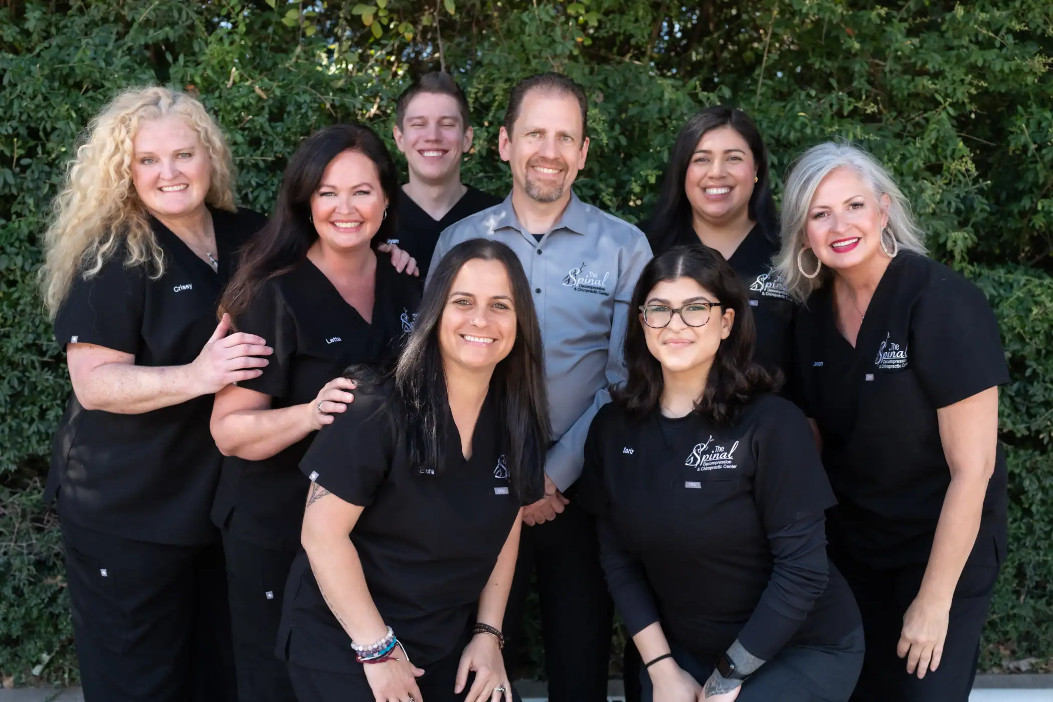 Eight adults in black medical scrubs, smiling and posing outdoors in leafy greenery. One person in the back row wears a light gray top. This friendly group of chiropractors offers spinal decompression services in Denton, Texas.