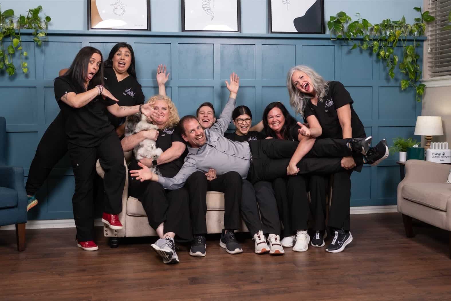 A group of seven smiling adults, including a local chiropractor in black uniforms, pose playfully on and around a beige couch in Denton, Texas. Two small dogs sit on laps. The background features blue paneling, plants, and framed art.