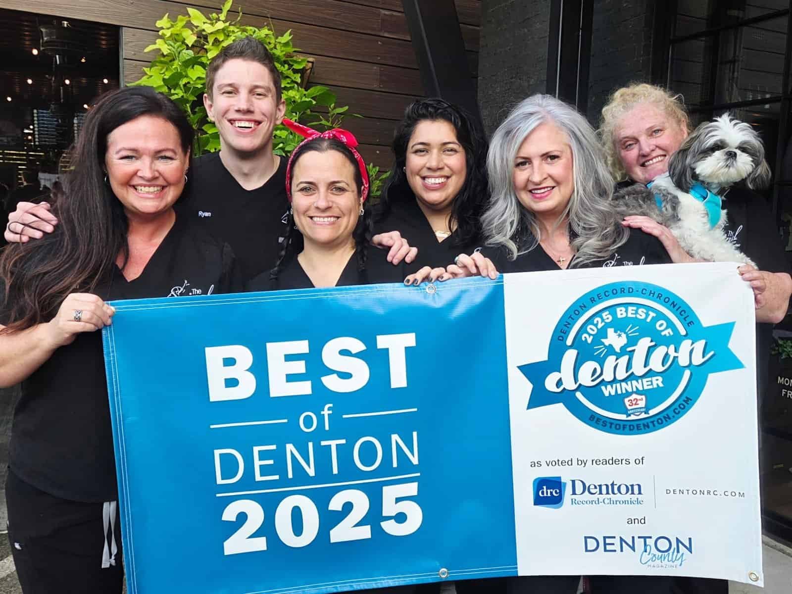 Six smiling people, including a chiropractor from Denton, Texas, stand outdoors holding two blue banners that read Best of Denton 2025 and display the 2025 Best of Denton Winner seal and local sponsor logos.