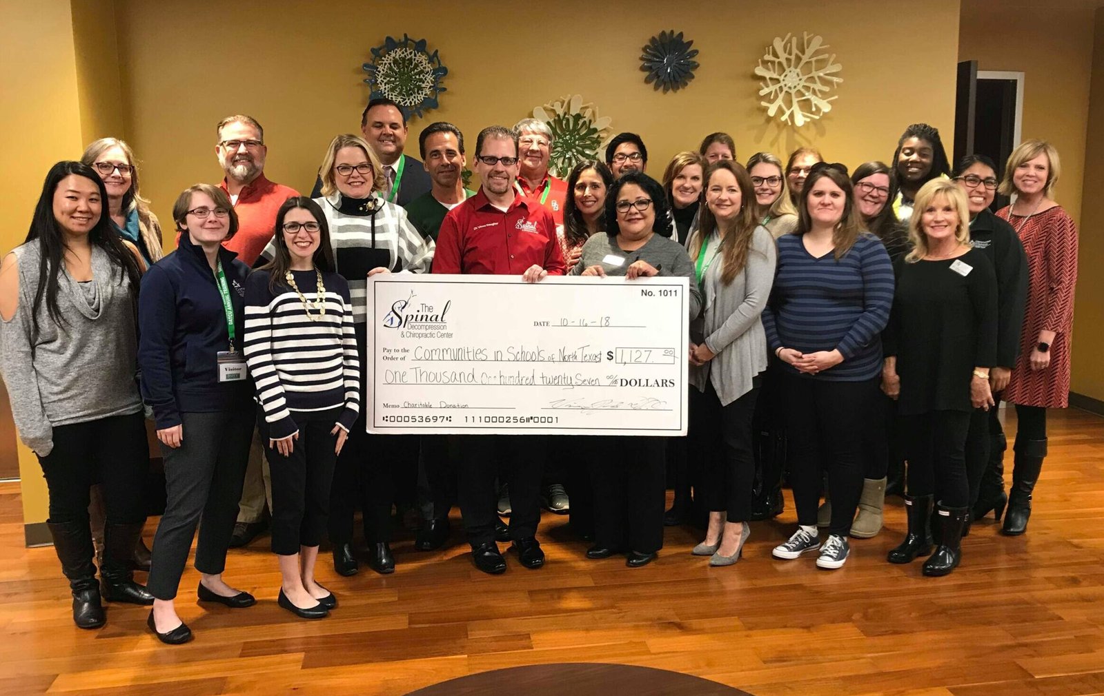 A group of people stands indoors smiling at the camera, with several holding a large ceremonial check for $1,187 made out to Community Partnership for Inclusive Living. A decorative wall piece and a local Denton Texas chiropractor are visible in the background.