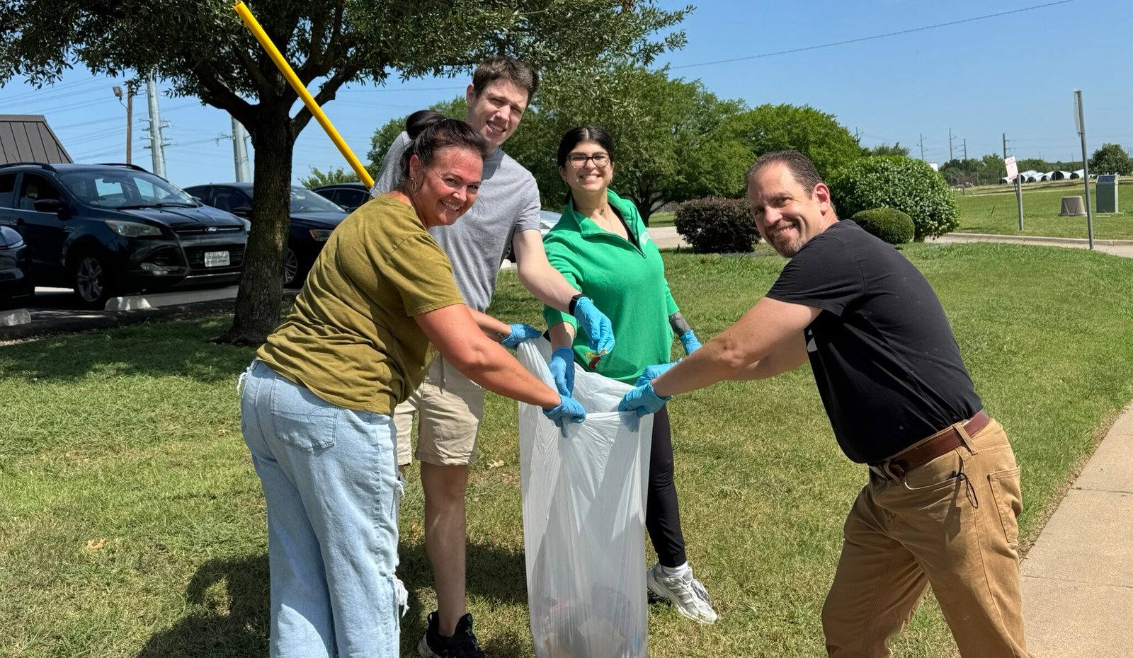 Four adults smiling and picking up litter together outdoors on a sunny day in Denton, Texas, placing trash into a large white bag. They are wearing gloves and standing on grass near parked cars and trees.