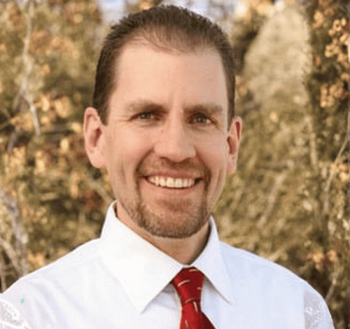 A man with short brown hair and a trimmed beard smiles outdoors in Denton, Texas, while wearing a white shirt and red patterned tie; trees with brownish-green foliage are visible in the background.
